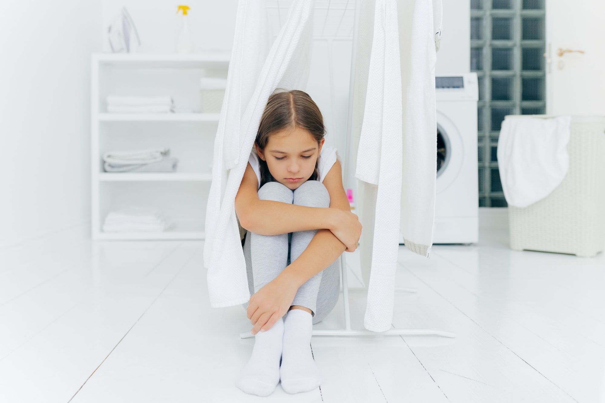 Lonely thoughtful sad small girl sits on floor in washing room near clothes dryer