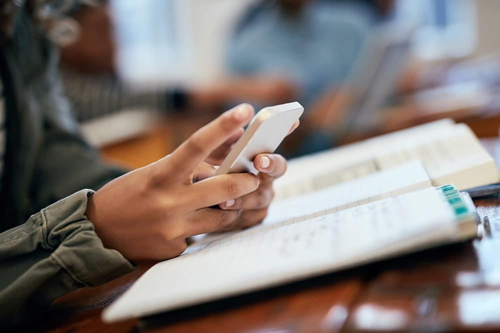 Twee handen van een tiener met een smartphone boven een schoolboek