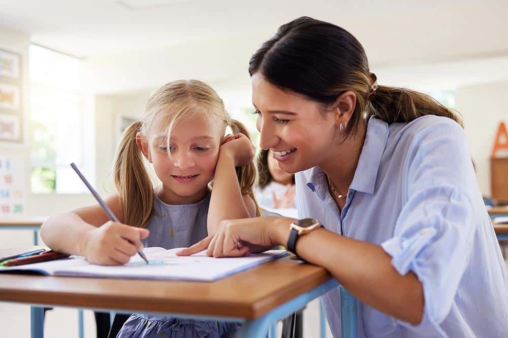 Een leerkracht zit gehurkt naast een leerling om haar te helpen met haar werk