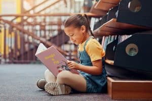 Een kind zit op de grond tegen een boekenkast aan een boek te lezen.