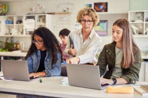 Docent begeleidt twee middelbare scholieren die op laptops werken.