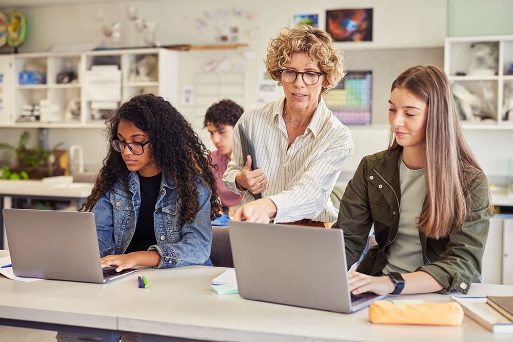 Docent begeleidt twee middelbare scholieren die op laptops werken.