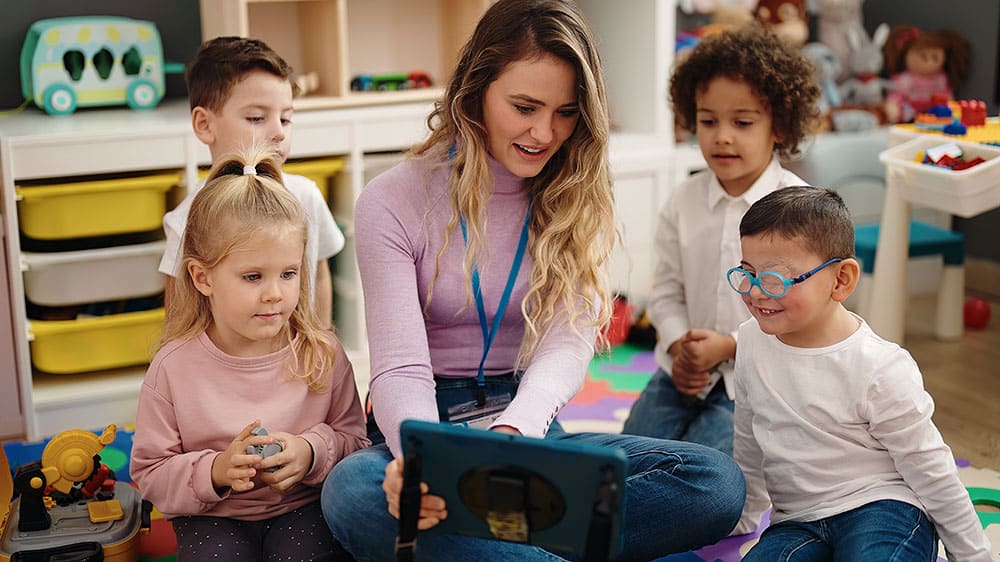 Leerkracht zit met een groep kleuters op de vloer van het klaslokaal en bekijkt samen met hen iets op een tablet.